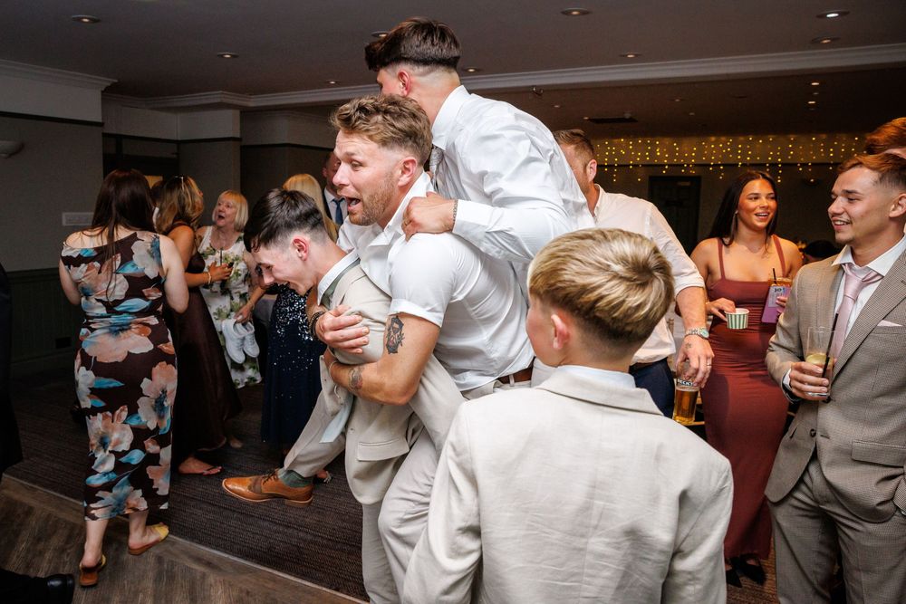 Guests celebrating at a wedding reception, with groomsmen playfully piggyback riding on the dance floor.