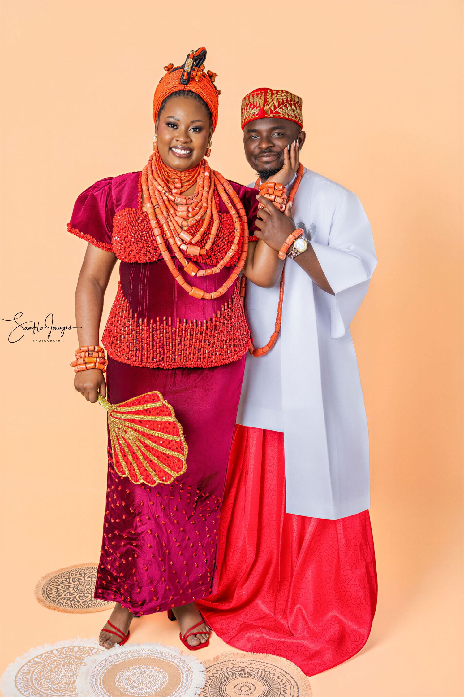 Couple in traditional Nigerian Urhobo attire with coral beads, woman in magenta and man in white agbada, posing for portrait.
