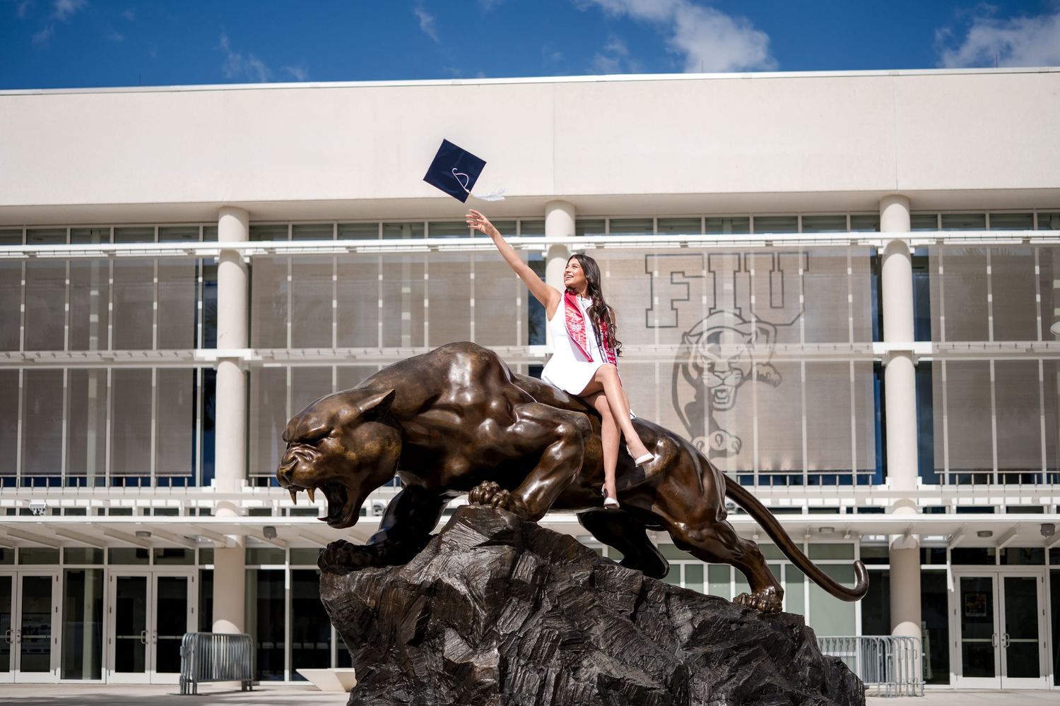 FIU graduate in cap and gown sitting atop the Panther statue, holding diploma, outside FIU arena.