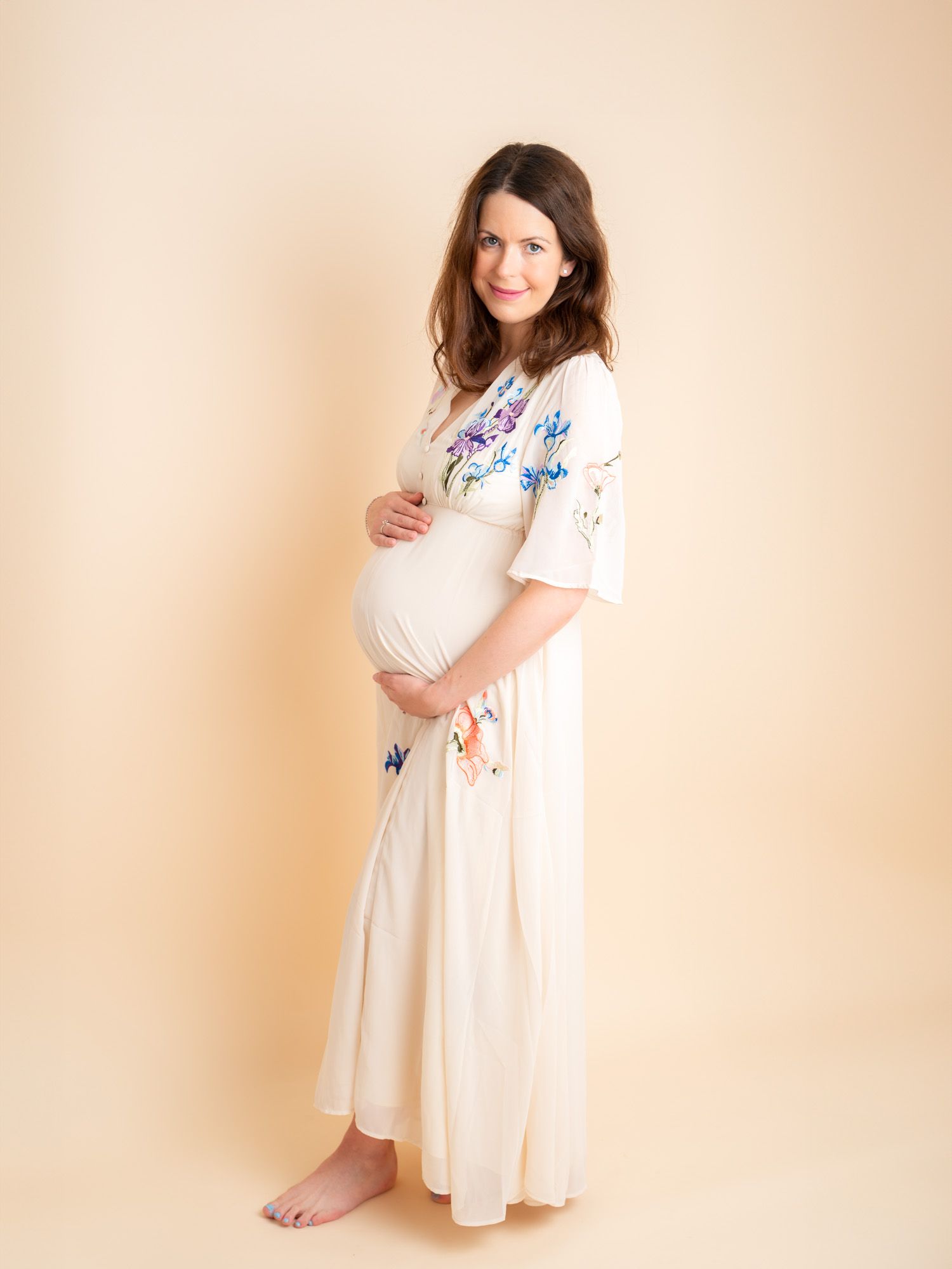 Pregnant woman in floral embroidered cream maxi dress cradling baby bump, barefoot on beige background.