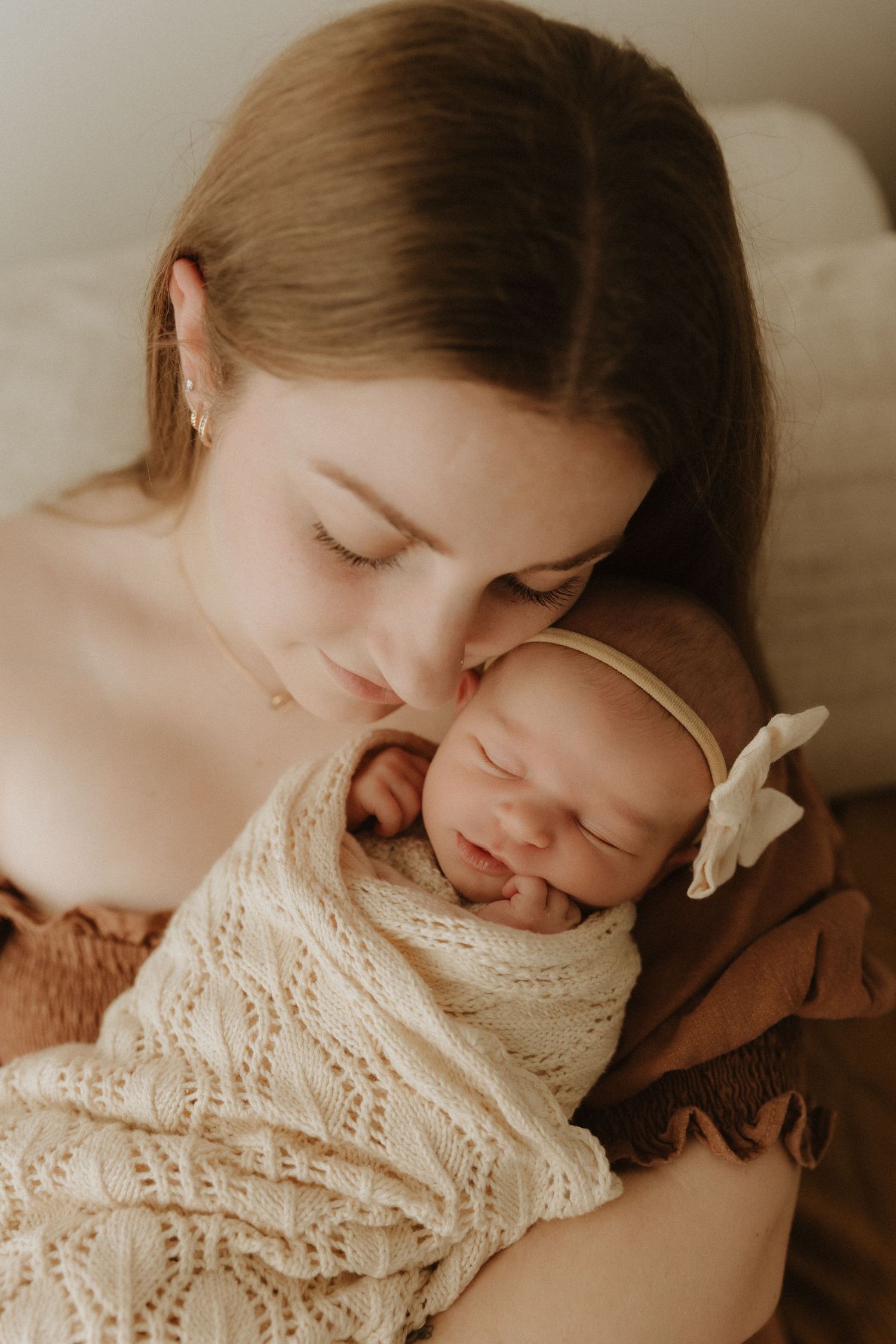 A young mother tenderly kisses her sleeping newborn baby wrapped in a cream knit swaddle with a white bow headband.