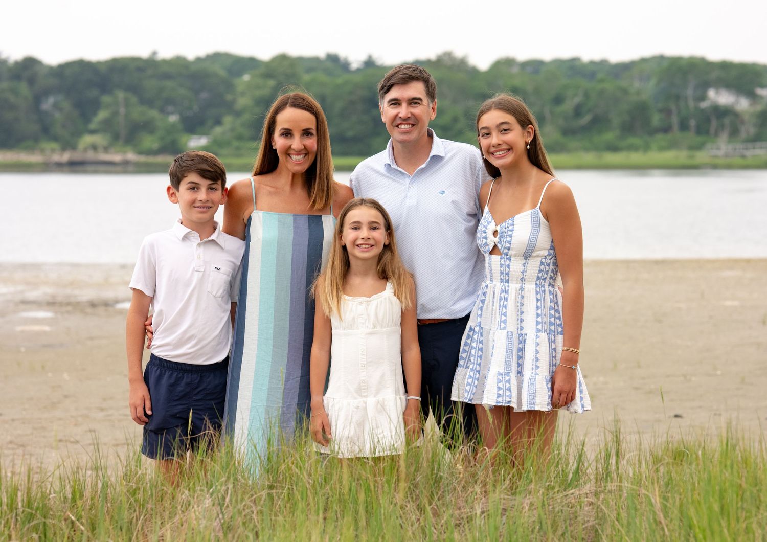 Happy family of five posing together on a sandy beach shore with a calm river and green trees in the background.