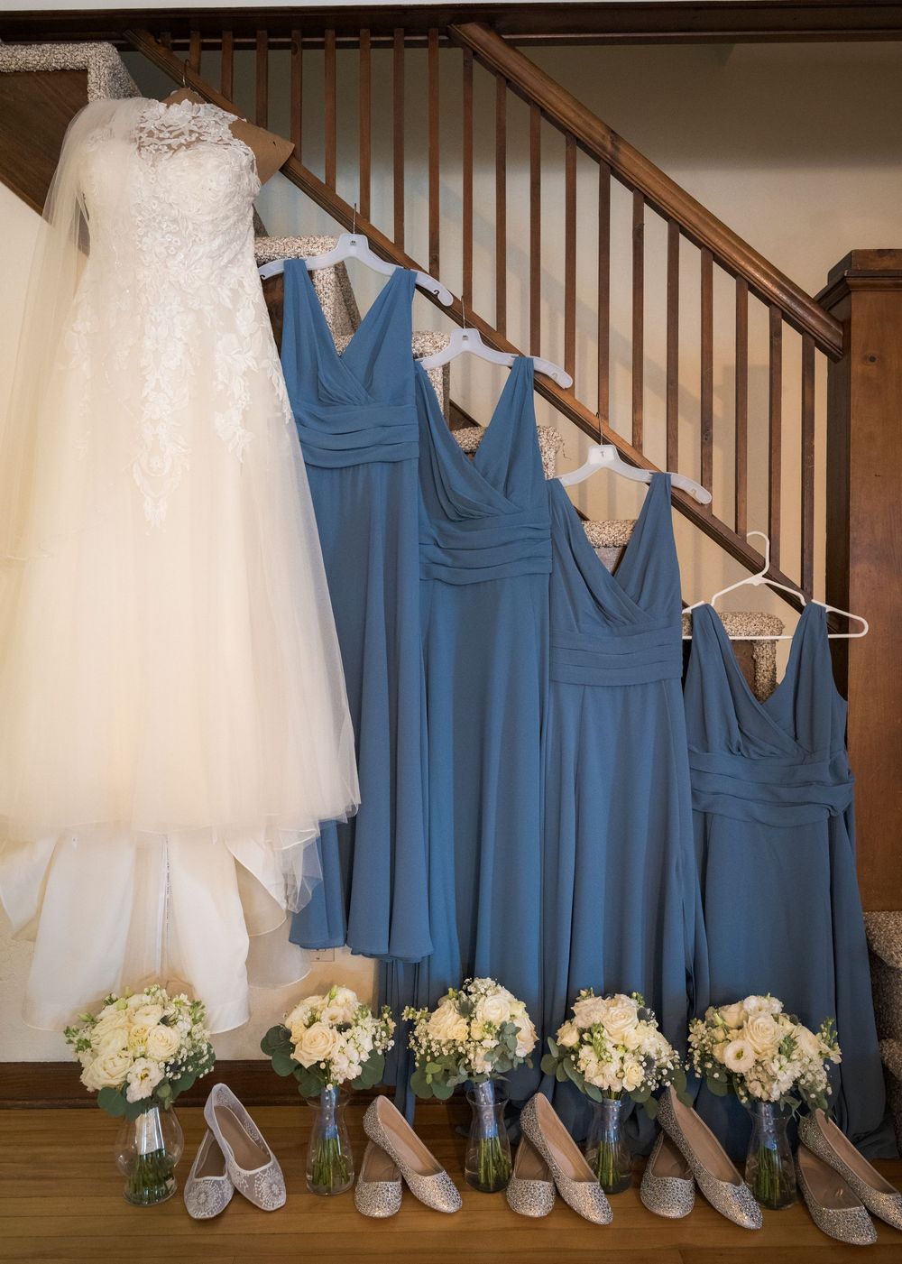 White wedding gown and blue bridesmaid dresses hanging on staircase with bouquets and heels below.