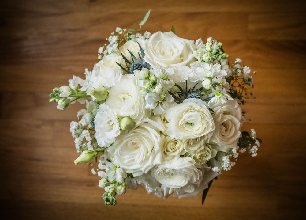 Elegant bridal bouquet of white roses and ranunculus with blue thistle accents on a wooden surface.