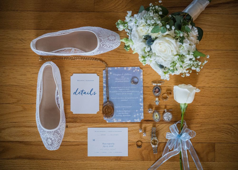 Elegant wedding flat lay featuring lace bridal shoes, blue invitations, white floral bouquet, and jewelry on wood floor.