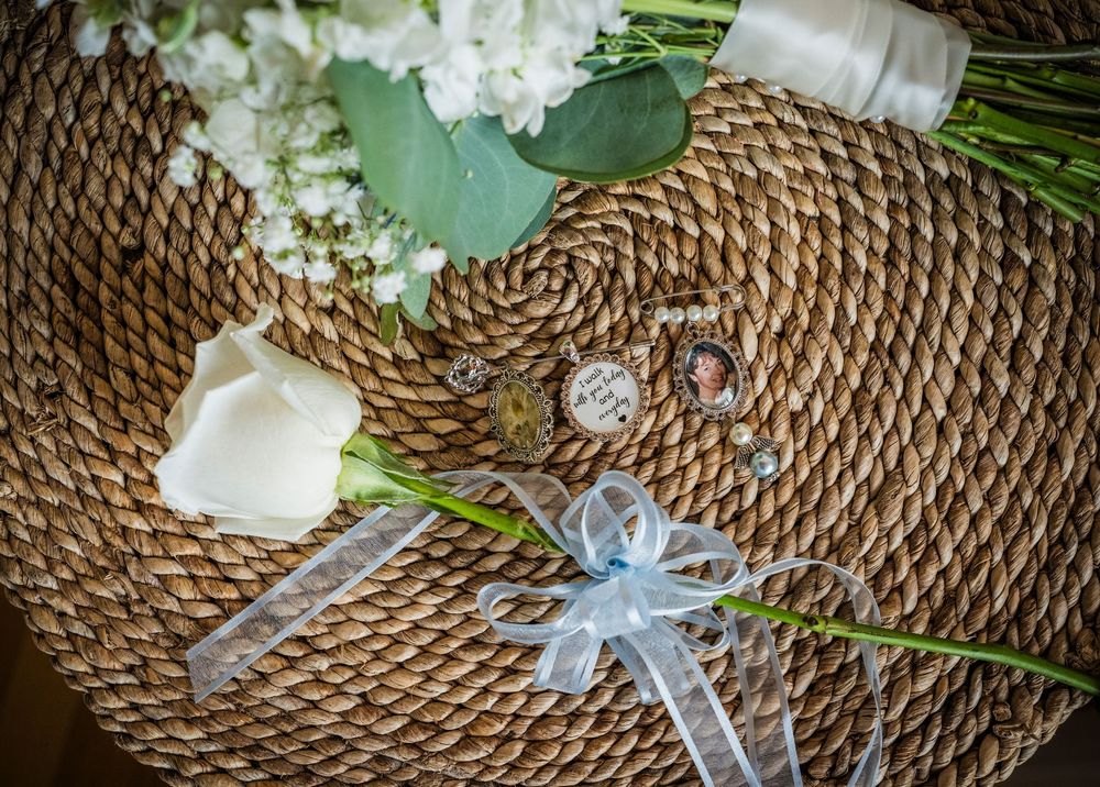 Wedding detail flat lay with white roses, boutonniere, and decorative locket on woven rattan surface.