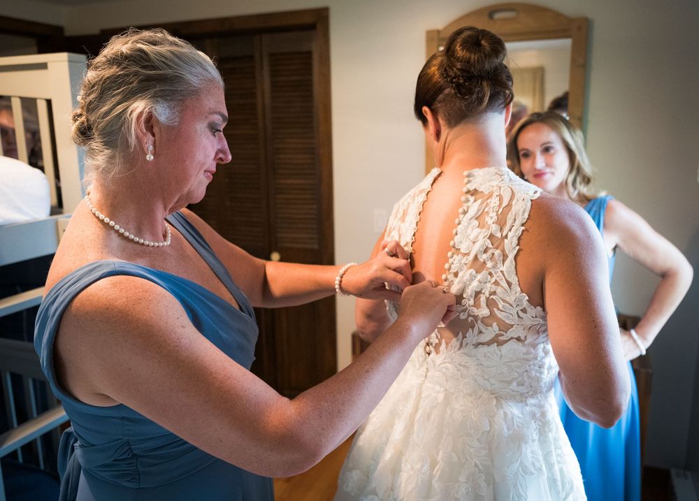 Mother helping bride fasten lace back wedding gown while bridesmaid watches in blue dress.