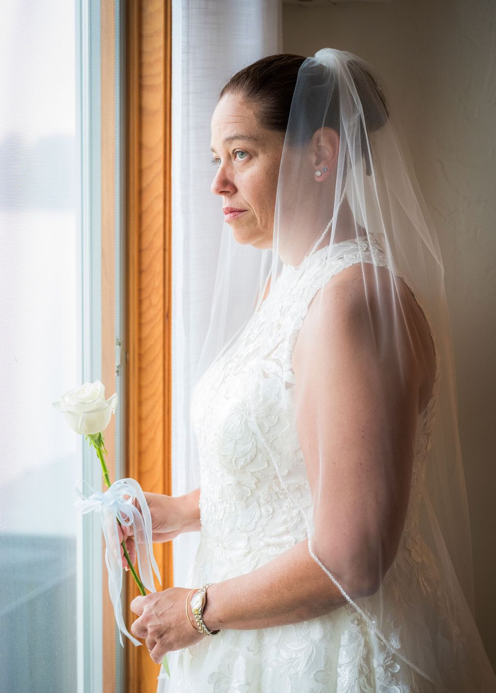 Bride in white lace gown and veil holds a rose, gazing thoughtfully out a window on her wedding day.