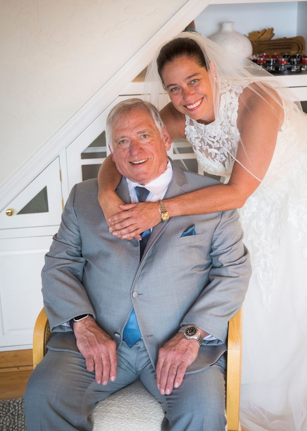 Bride in lace veil embraces seated older man in gray suit indoors before wedding ceremony.