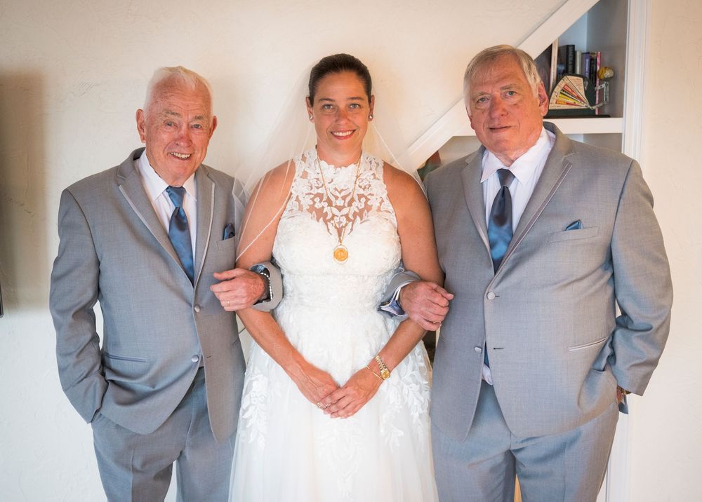 Bride in white lace dress stands between two men in grey suits with blue ties at a wedding celebration.