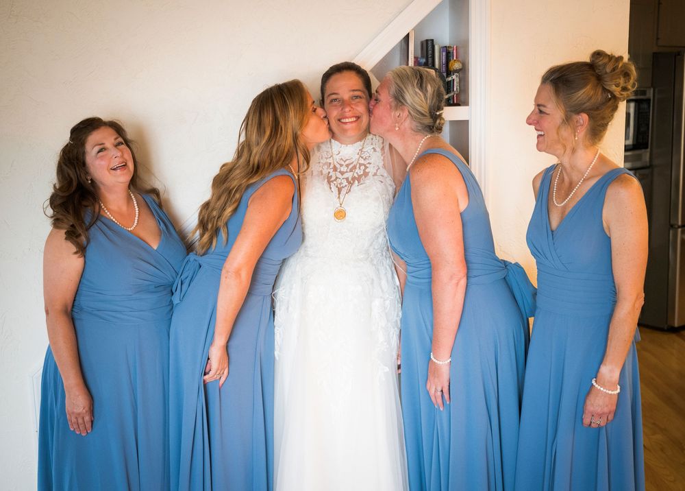 Bride in white lace gown receives kisses from bridesmaids in blue dresses during wedding day preparations.