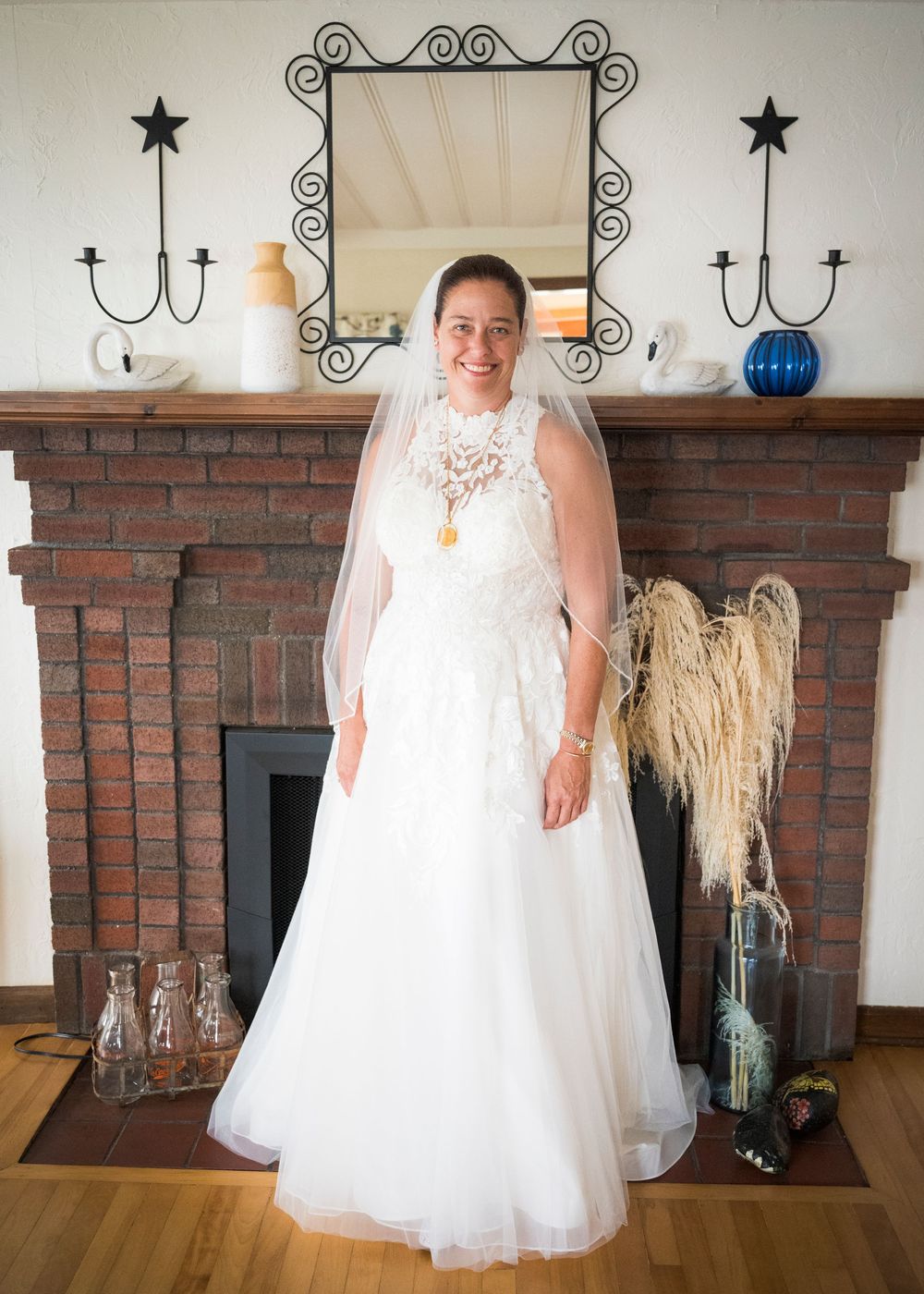 Smiling bride in lace halter wedding gown and veil stands before brick fireplace mantel.