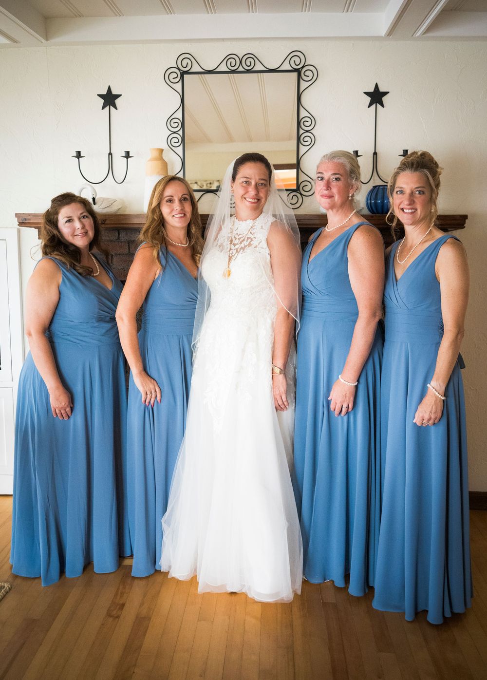 Bride in lace gown poses with four bridesmaids in matching steel blue dresses before fireplace.