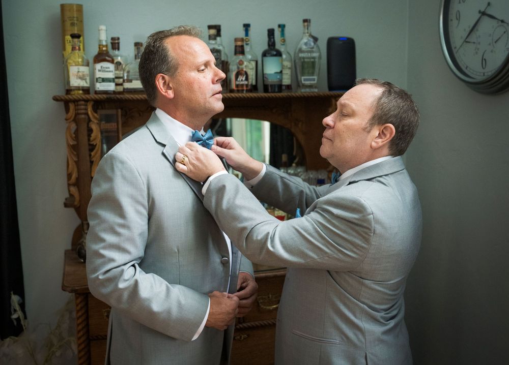 Two men in light gray suits, one adjusting the other's bow tie indoors near a bar shelf.