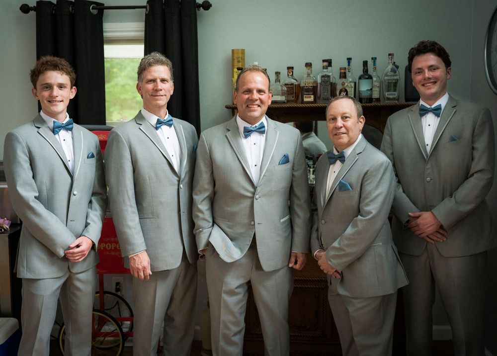 Five groomsmen in matching gray suits and blue bow ties posing together indoors before wedding.