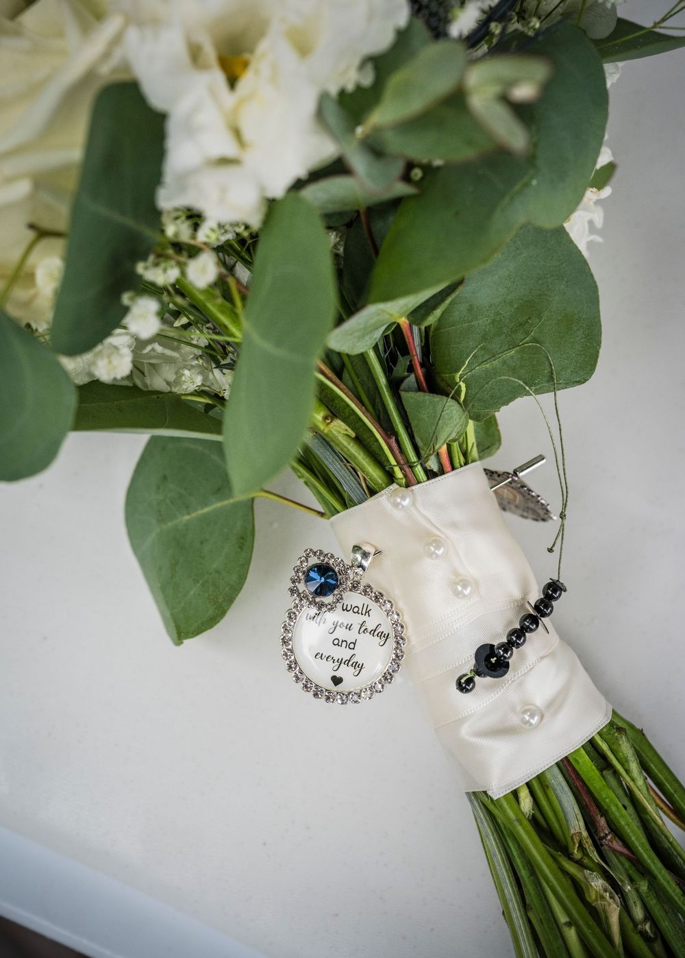 Bridal bouquet with white flowers and eucalyptus, adorned with pearl wrap, feather pin, and memorial charm pendant.