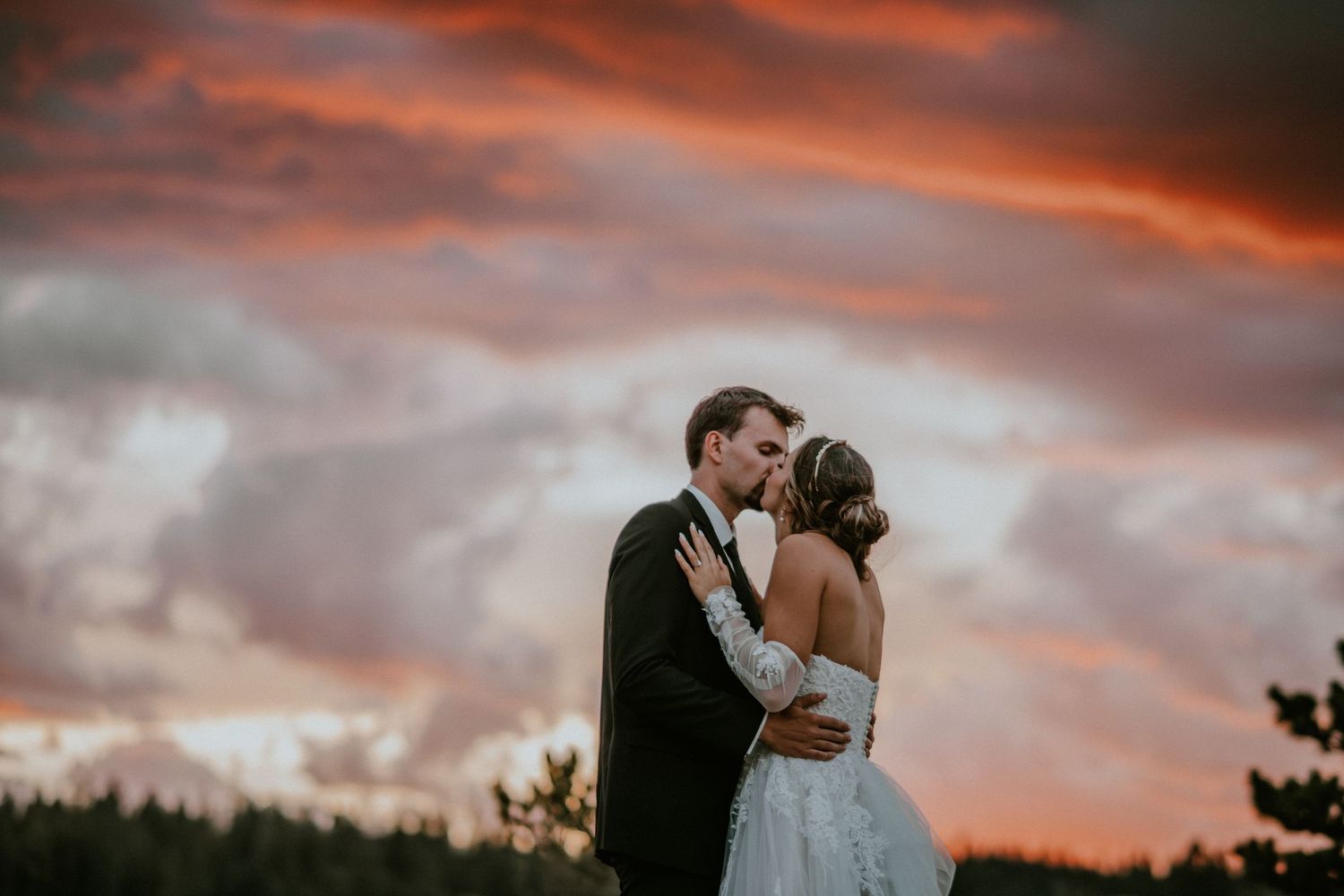 Bride and groom share a kiss at sunset with dramatic red and orange clouds over a forest backdrop.