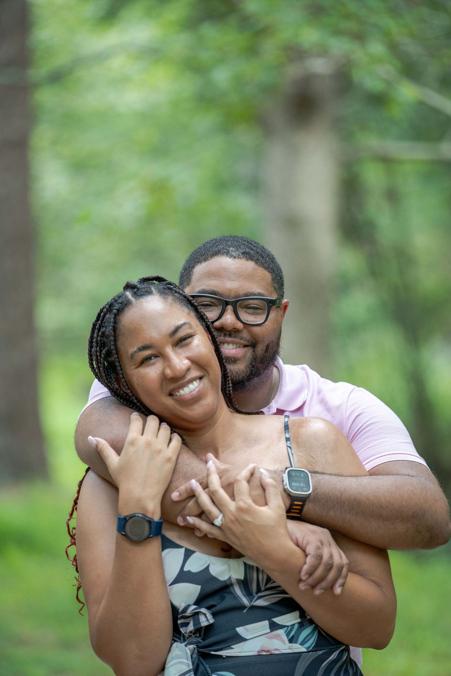 A joyful couple embraces outdoors in a lush green park, woman smiling brightly as man hugs her from behind.