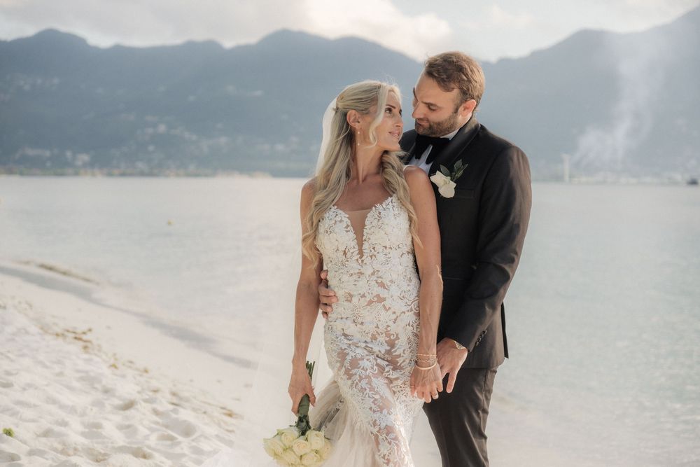 Bride and groom share a romantic moment on a sandy beach with mountains and water in the background.