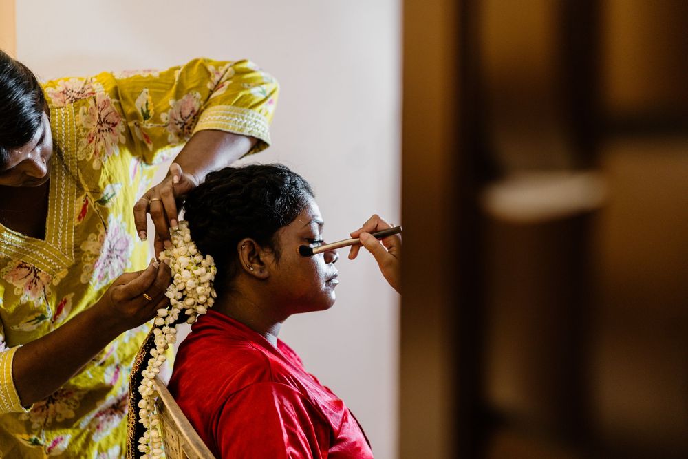 A bride in red gets makeup applied while someone arranges floral hair accessories during a traditional Indian wedding preparation.