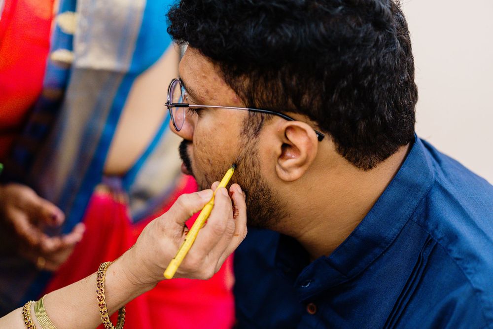 A man wearing glasses gets yellow face paint applied by a hand adorned with a gold bracelet during a cultural event.