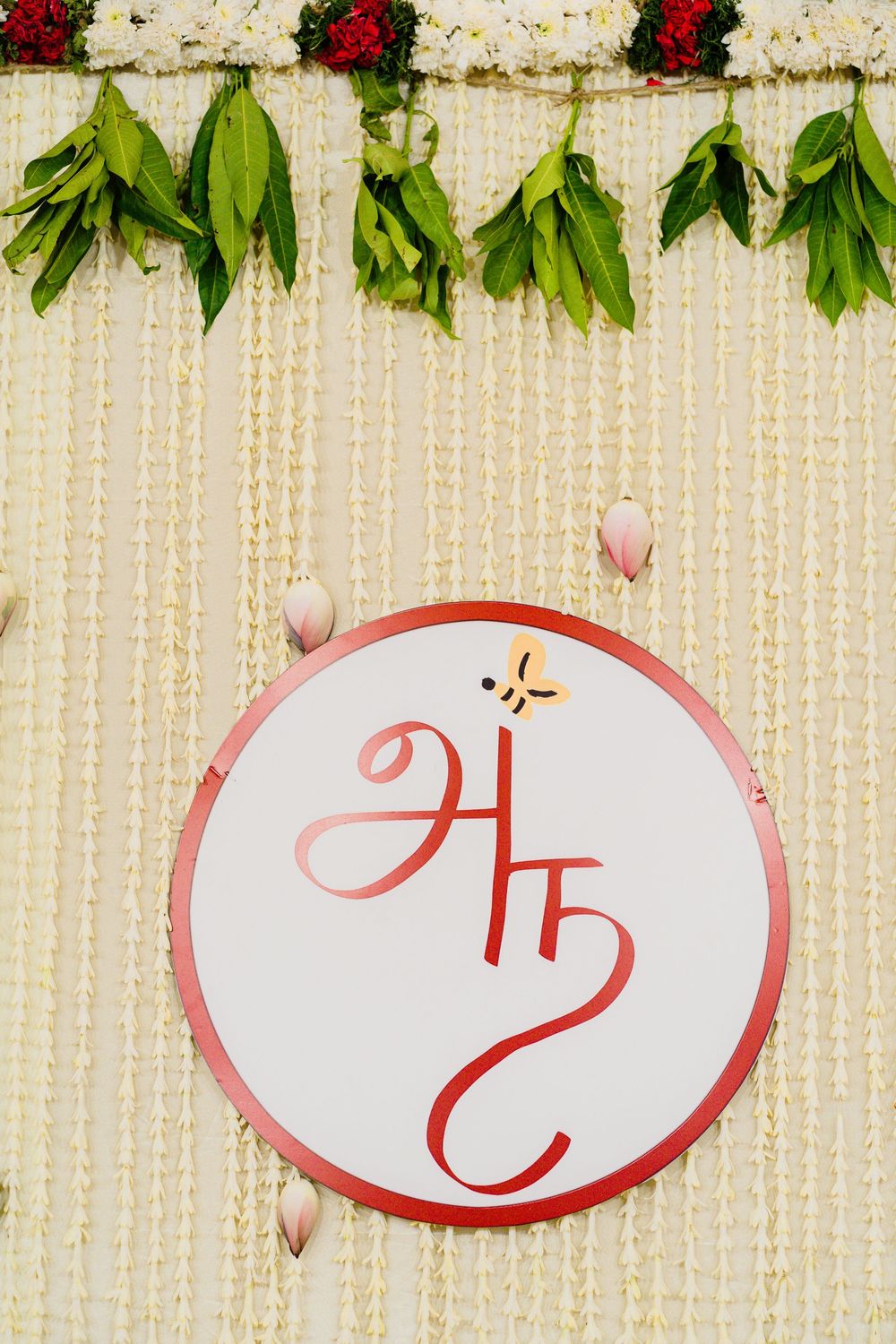 Decorative Indian wedding monogram sign with floral garlands featuring initials in red script on white circular backdrop.