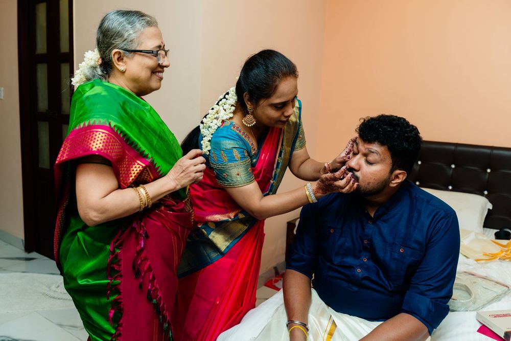 Two women in colorful sarees applying tilak to a young man during a traditional South Indian ceremony.