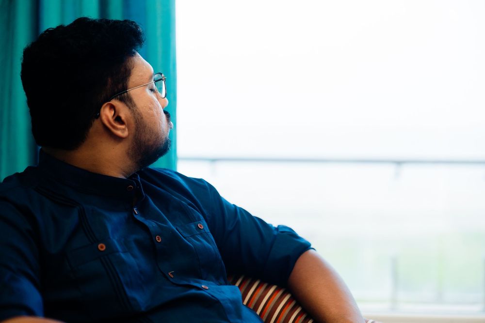 A thoughtful young man with glasses and a beard sits in a chair, wearing a blue shirt, gazing away in a serene setting.