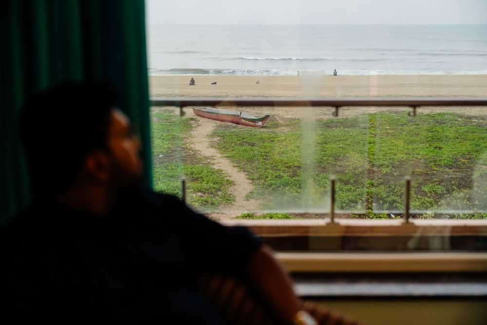 Man gazing out train window at scenic beach with boats and ocean waves visible through glass.