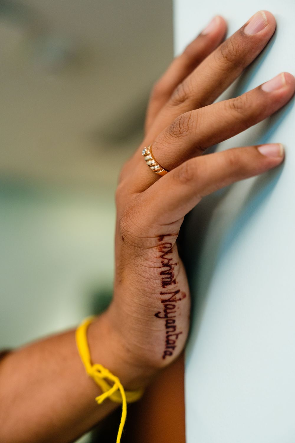 Close-up of a hand with henna tattoo writing and gold ring, resting against a white wall with yellow thread bracelet.