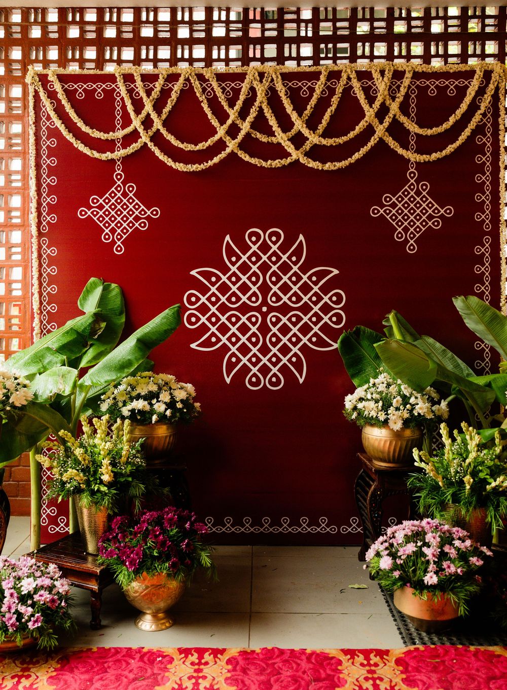 Traditional Indian kolam art on red backdrop with marigold garlands, brass pots, and tropical plants for festive decor.