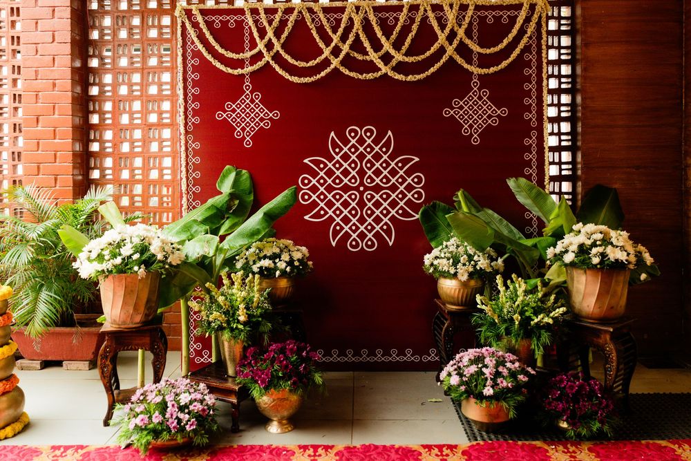 Traditional South Indian wedding altar with kolam rangoli, banana leaves, marigold flowers, and brass pots on red backdrop.