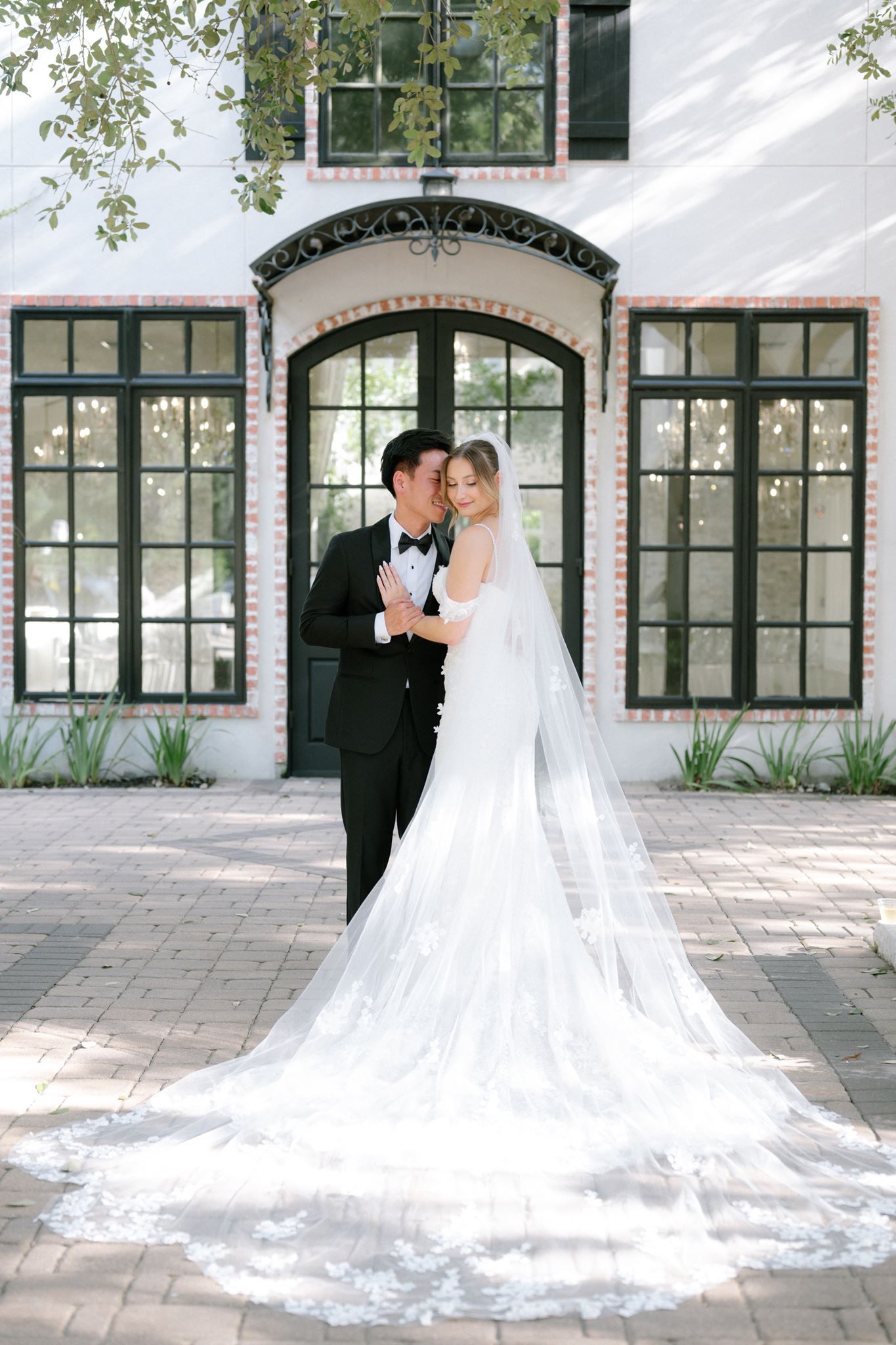 Bride in lace gown with cathedral veil embraced by groom in black tuxedo outside elegant white building.