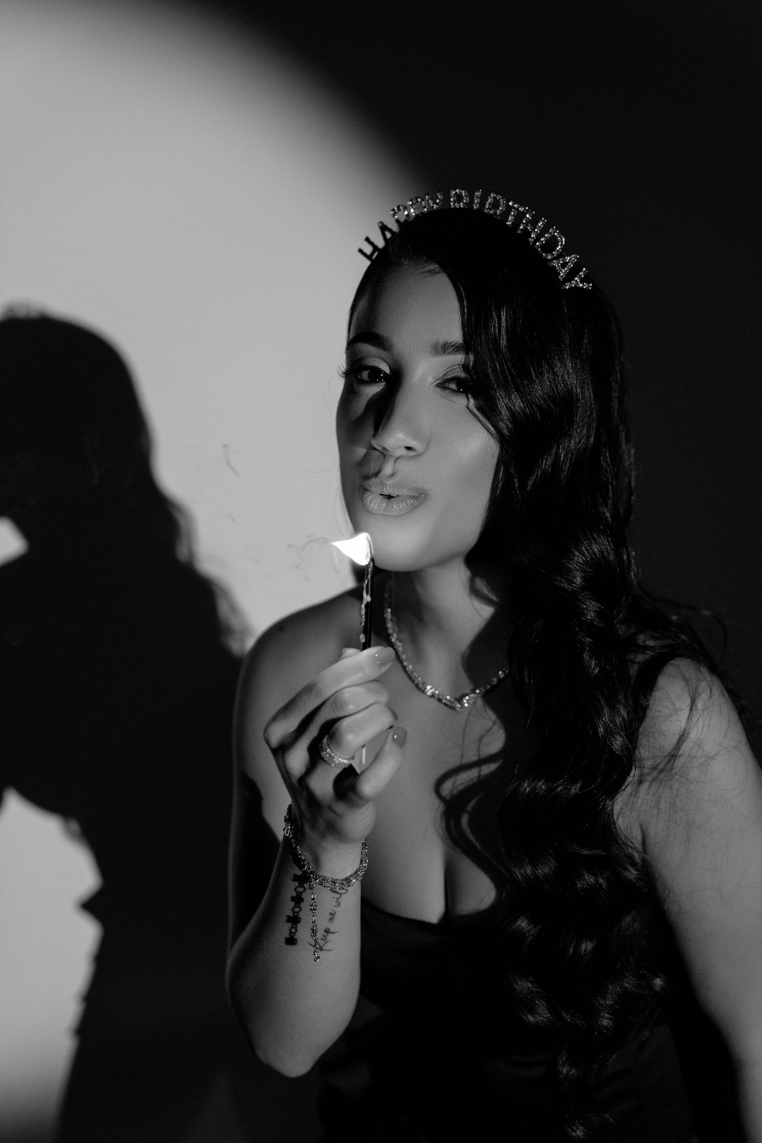 Black and white portrait of a woman wearing a Happy Birthday tiara, blowing out a candle, with shadow on wall.