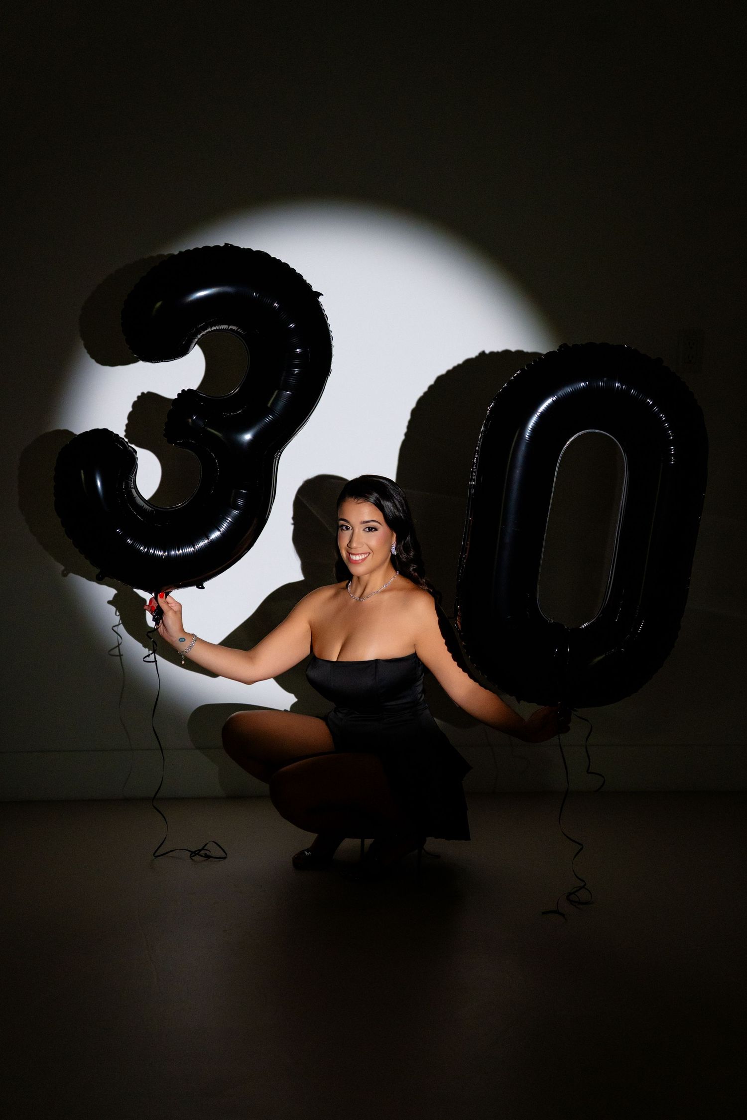 Woman in black dress crouching with large black number 30 balloons celebrating her 30th birthday.