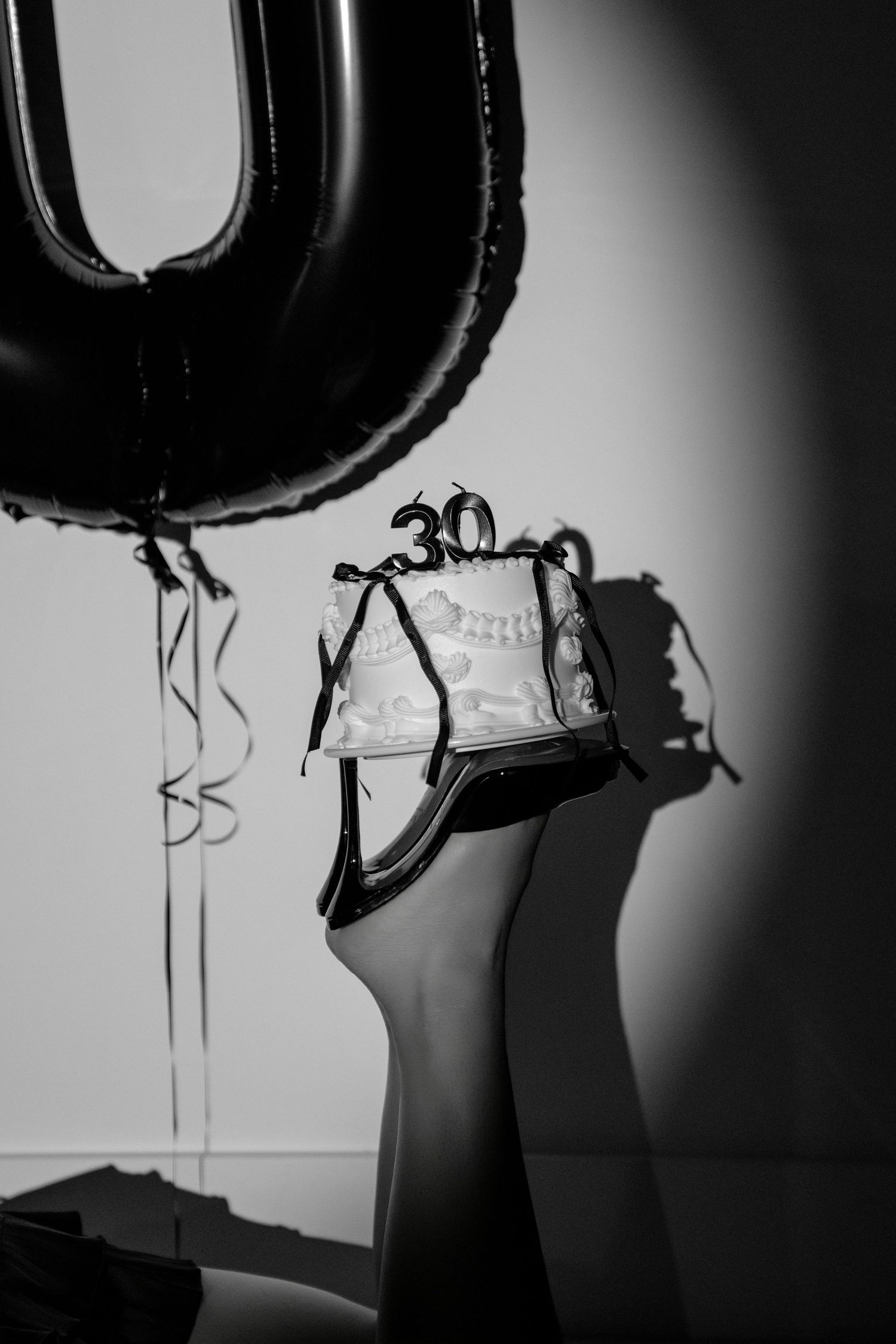 Black and white photo of a 30th birthday cake balanced on a heel with number balloons in the background.