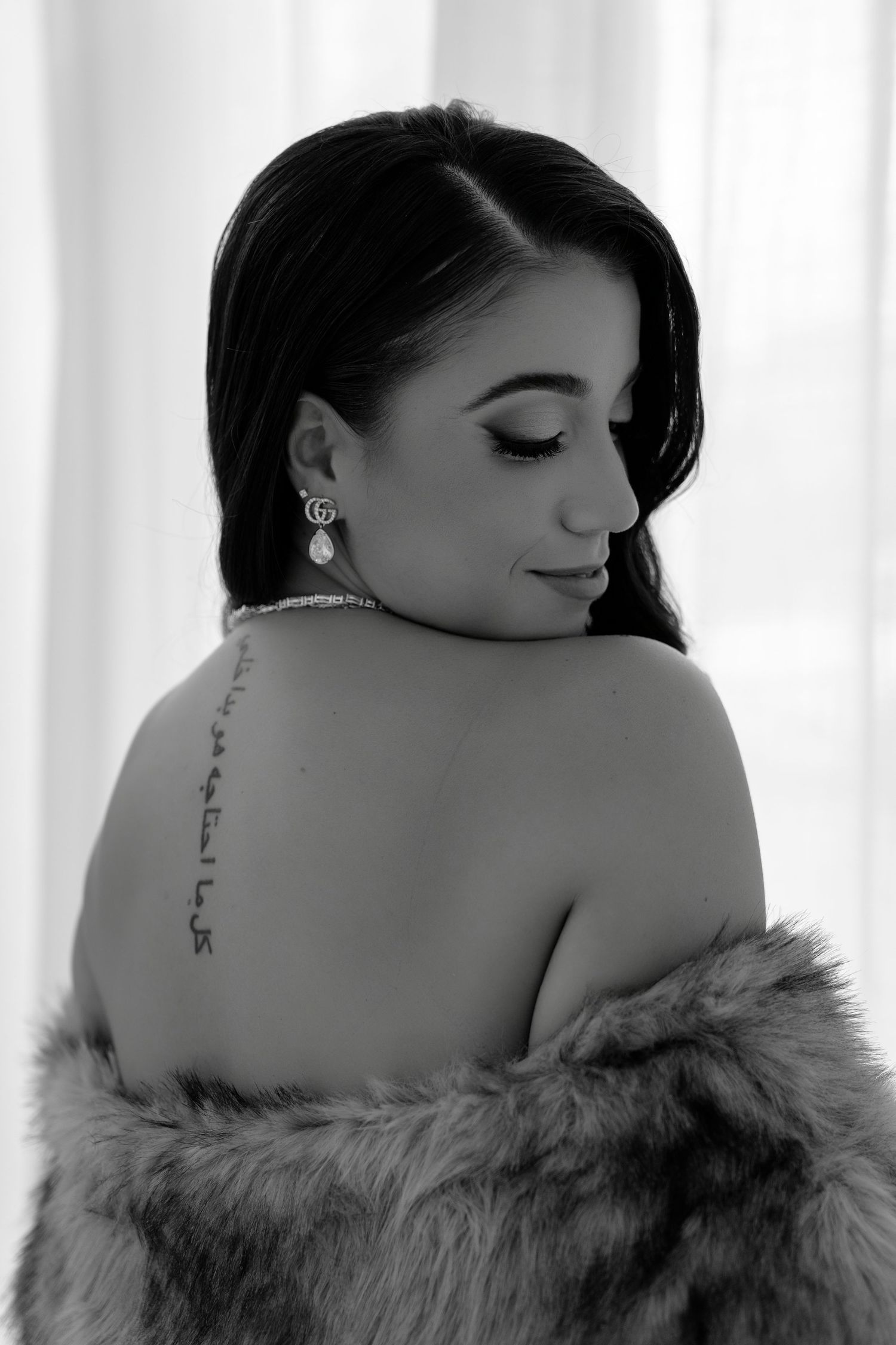 Black and white portrait of a woman with a spine tattoo, wearing a fur wrap and elegant earrings, looking over her shoulder.