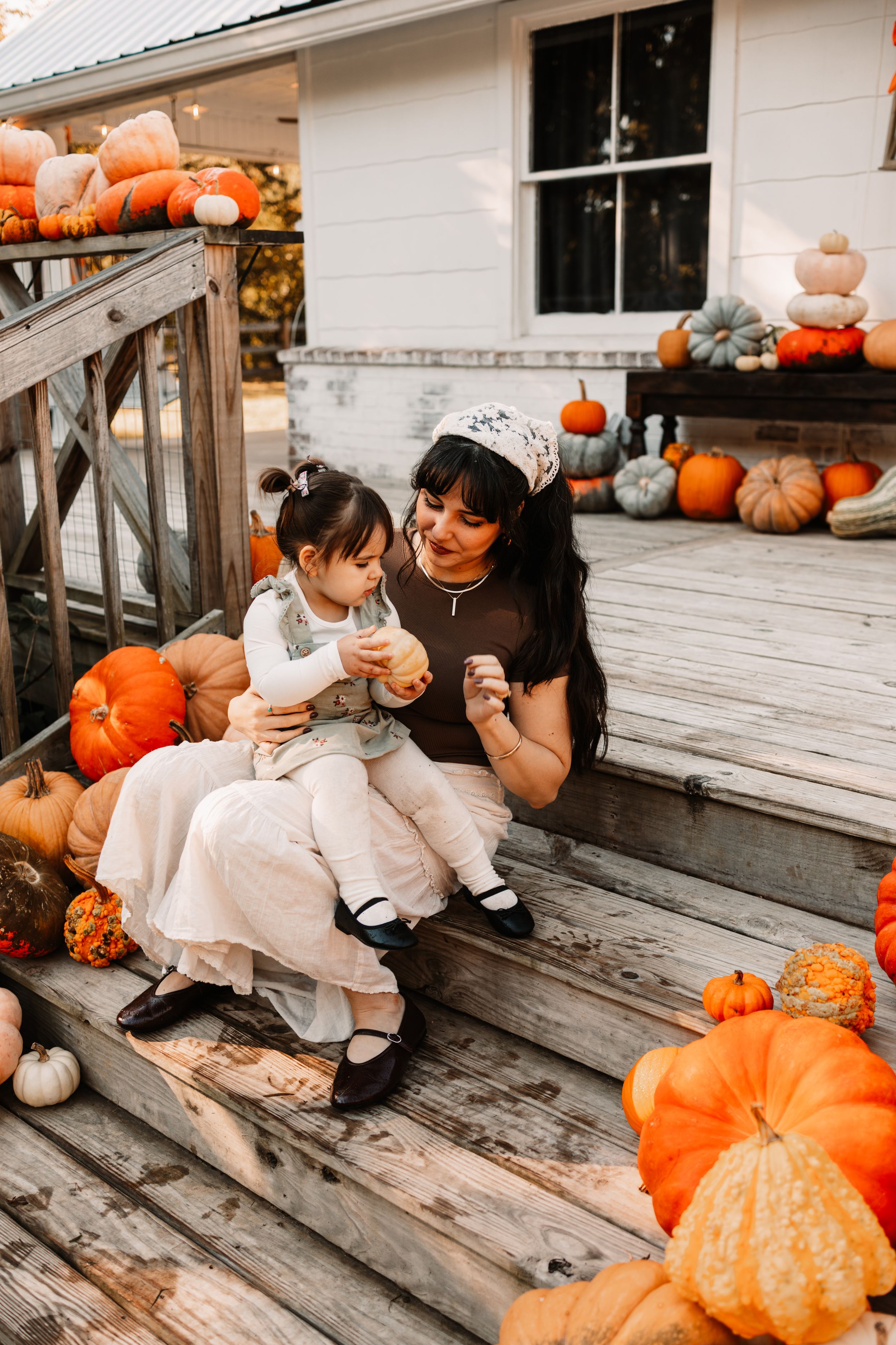 A mother and toddler sit on wooden porch steps surrounded by colorful pumpkins during autumn at The Holmstead Studio in Karns, TN.