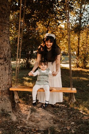 A smiling mother stands behind her toddler sitting on a wooden rope swing at The Holmstead Studio in Karns, TN with Nina Thomas Photography.