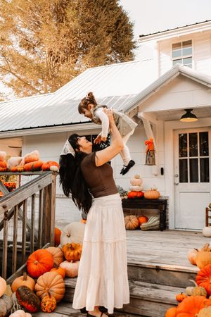 A woman lifts a child surrounded by pumpkins on a farmhouse porch in a festive fall autumn scene at The Holmstead Studio in Karns, TN.