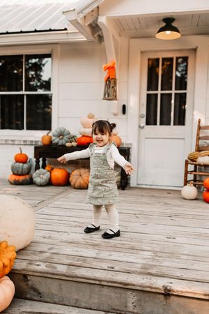 Toddler girl in green jumper dress stands on rustic porch surrounded by colorful pumpkins at The Holmstead Studio in Karns, TN.
