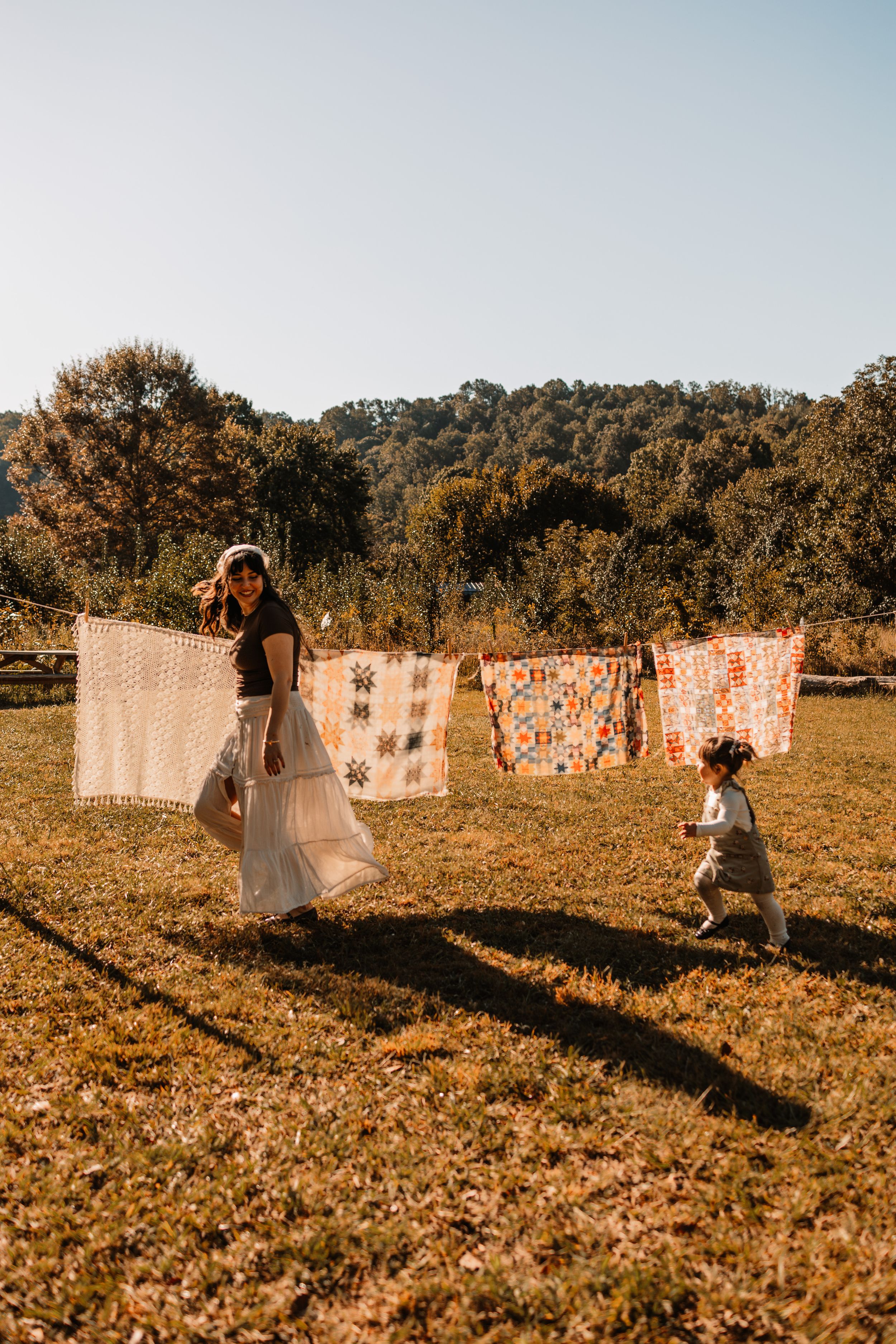 A woman in a white skirt and a toddler play outdoors near colorful quilts at The Holmstead Studio in Karns, TN with Nina Thomas Photography.