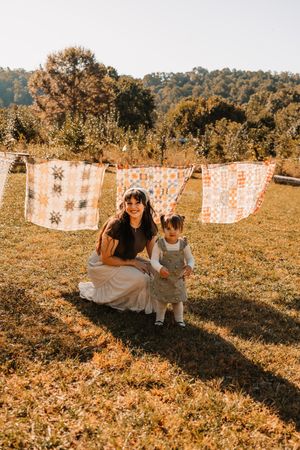 Mother crouching with toddler on sunny lawn with vintage quilts hanging on a clothesline in autumn at The Holmstead Studio in Karns, TN.