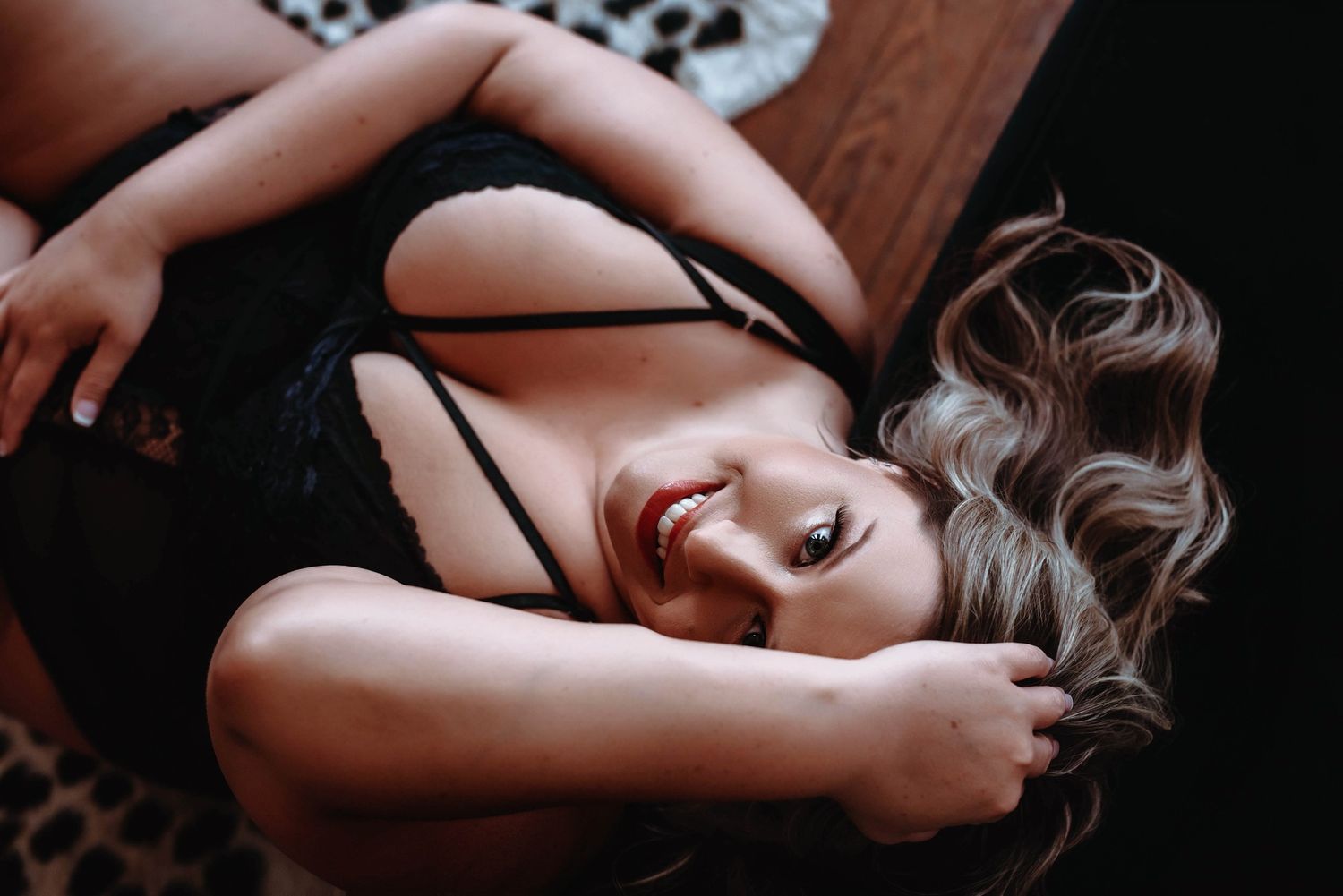 Woman in black lace lingerie lying down, smiling with wavy blonde hair in a dark, moody boudoir setting.