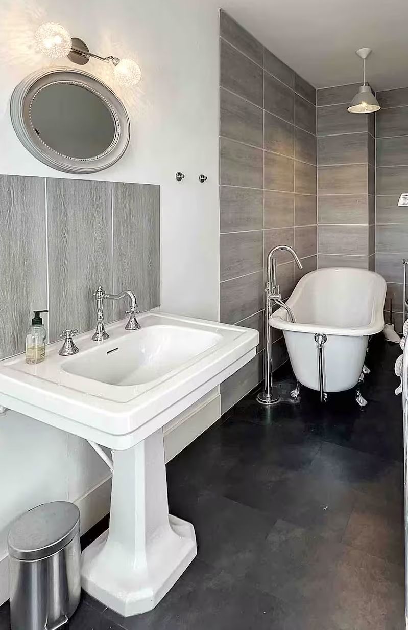 Rustic bathroom with exposed brick wall, round mirror, wooden vanity, and black vessel sink on hardwood floors.