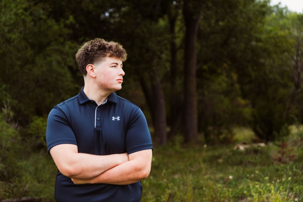 Young man in navy Under Armour polo stands with arms crossed outdoors among lush green trees.