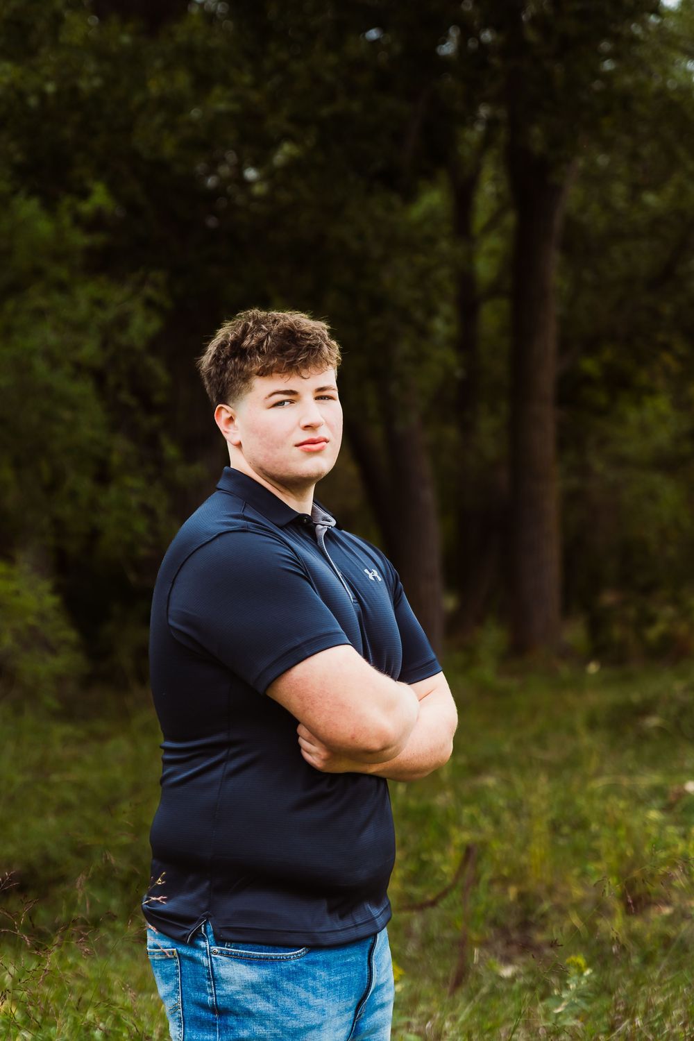 Young man in navy polo shirt stands with arms crossed in a wooded outdoor setting for senior portrait.