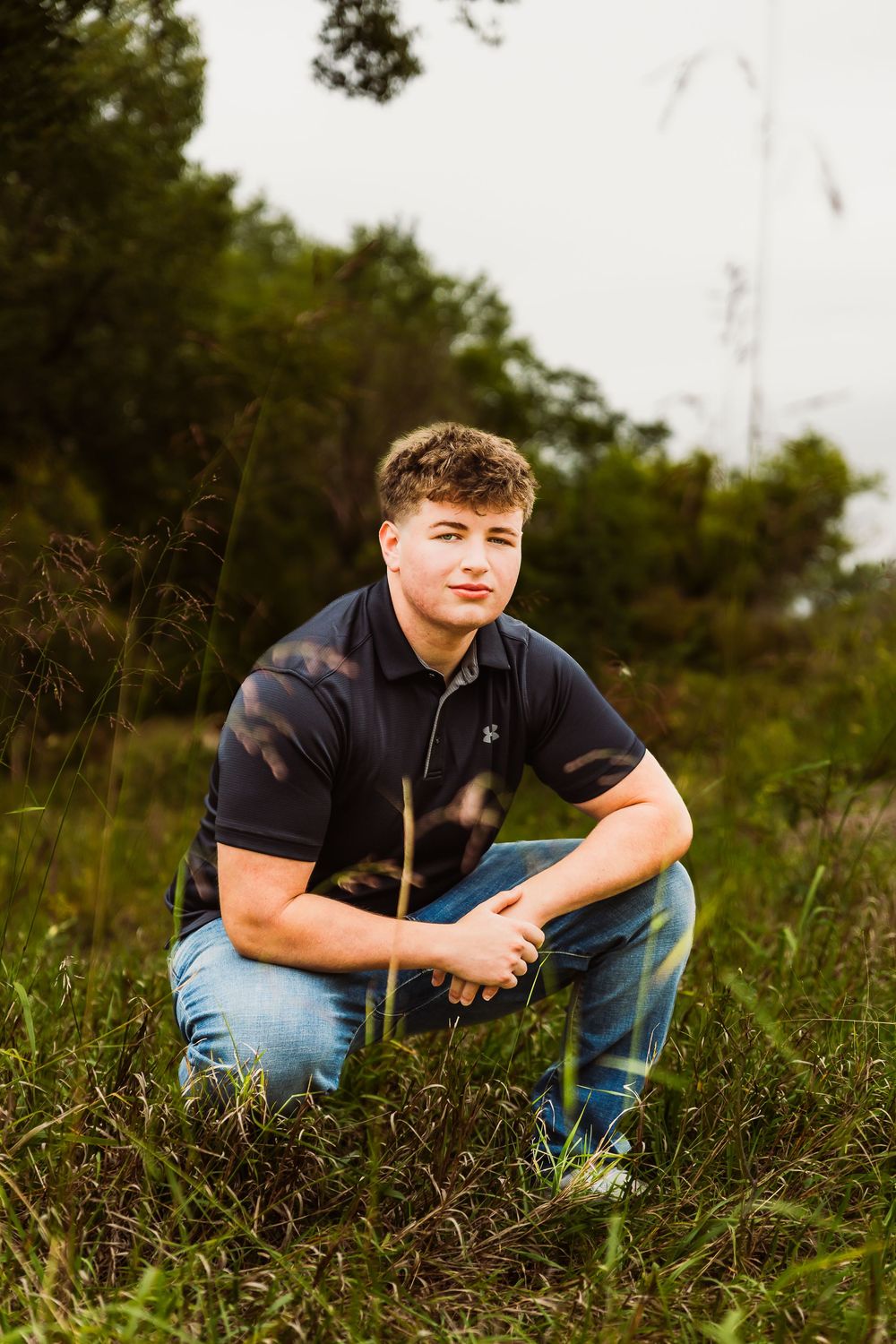 Young man in navy polo and jeans crouching in a grassy field with trees in the background.