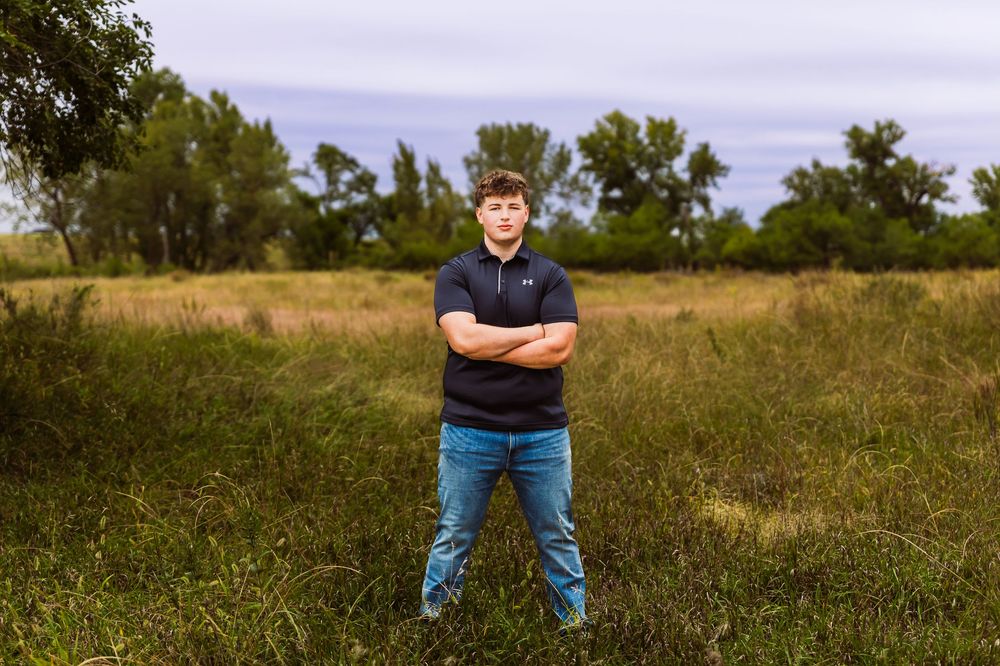 Young man standing confidently with arms crossed in a lush green field with trees in the background.
