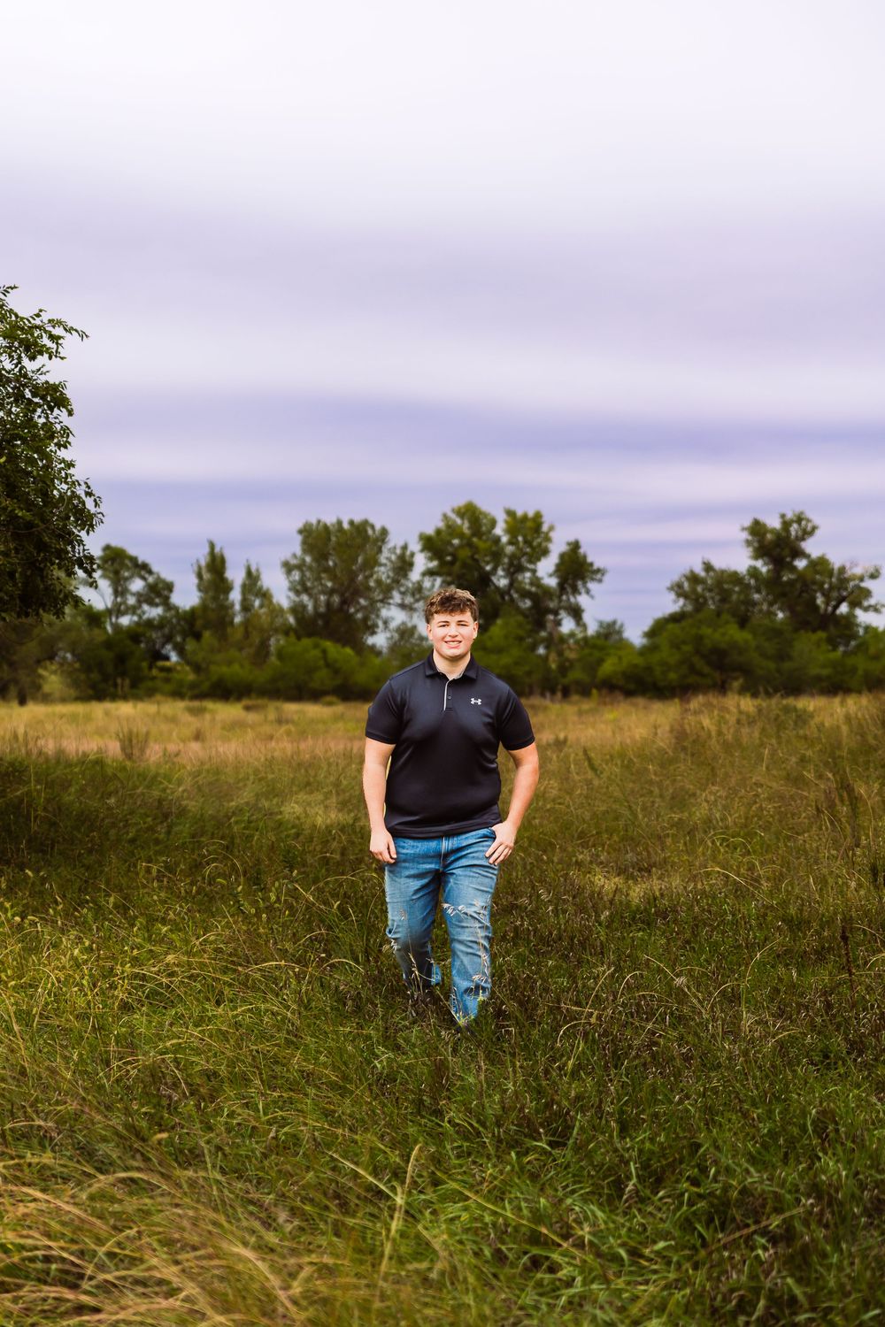 Young man in black polo and jeans walks through a lush green field with trees and cloudy sky behind him.